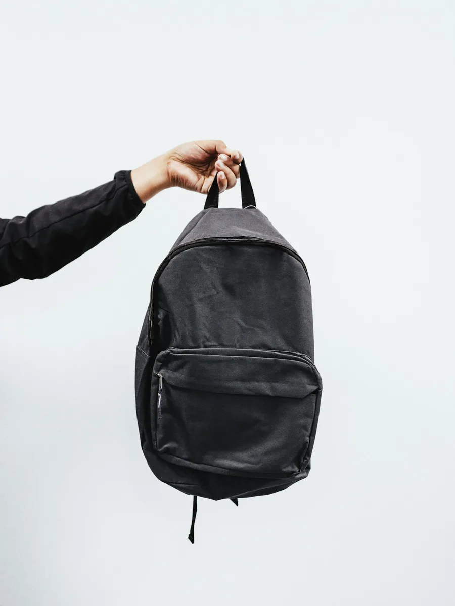 Une main tient un sac à dos noir devant un mur clair.