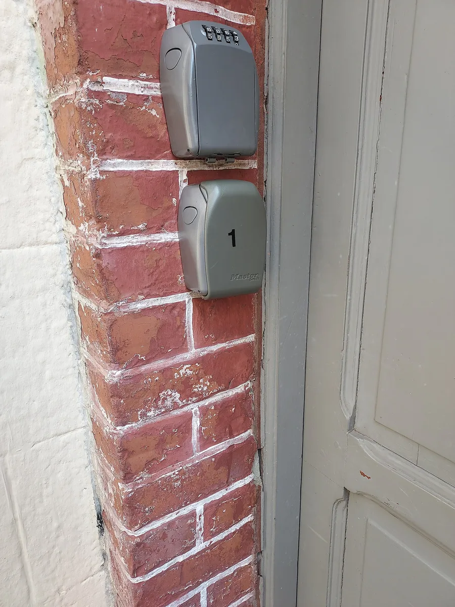 Deux boîtes à clés grises fixées sur un pilier en briques, à côté de la porte d'un immeuble.