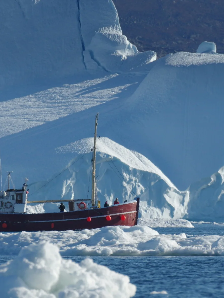 Un bateau de pêche rouge navigue devant un immense iceberg