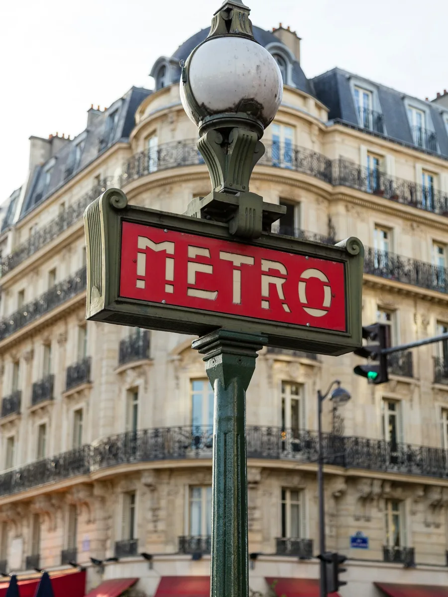 Enseigne rouge « Métro » surmontée d'un lampadaire, devant un immeuble haussmannien, place de la Bastille à Paris