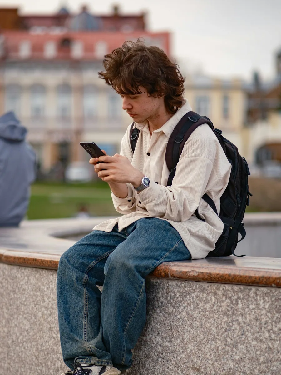 Un jeune homme avec un sac à dos, assis sur un muret, regarde son téléphone