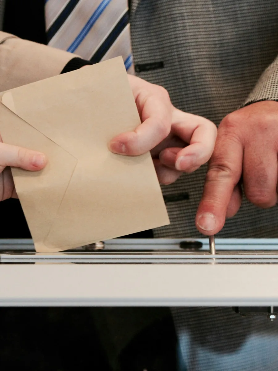Des mains glissent une enveloppe de vote marron vers la fente d'une urne