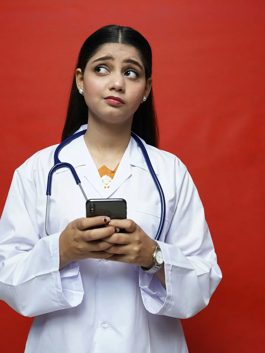 Une femme médecin en blouse blanche, stéthoscope autour du cou, consulte son smartphone d'un air perplexe