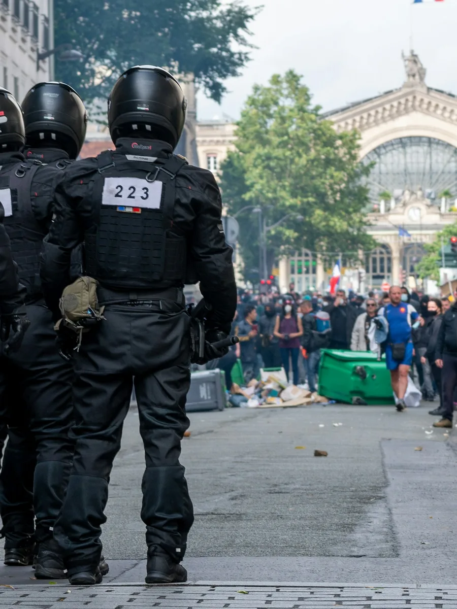 Des policiers casqués en tenue de maintien de l'ordre, vus de dos, font face à une foule de manifestants devant la gare du Nord à Paris