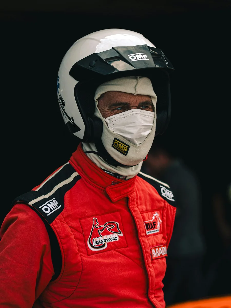 Membre d’une équipe de course en combinaison rouge et casque blanc, le visage couvert d’un masque, au bord d’un circuit.