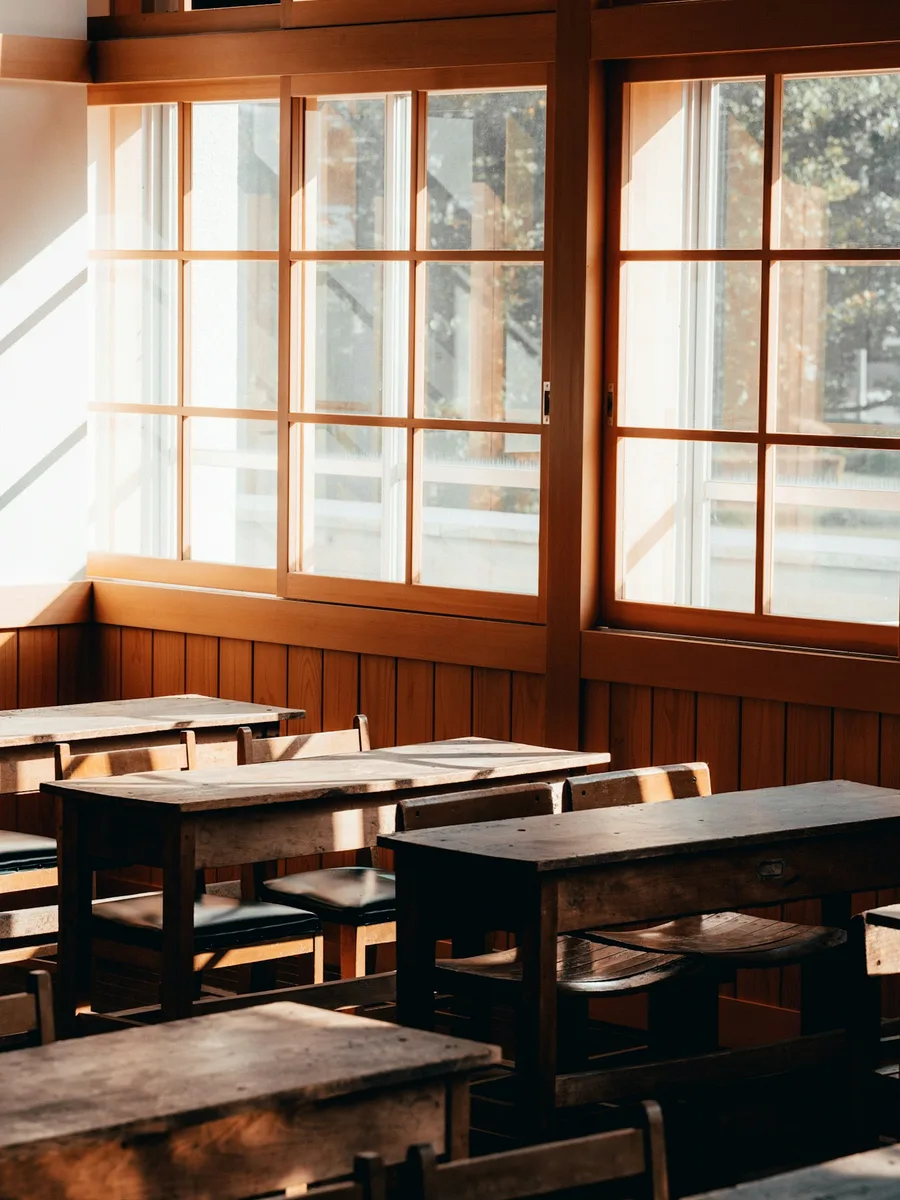 Une salle de classe vide aux pupitres en bois, éclairée par de grandes fenêtres.