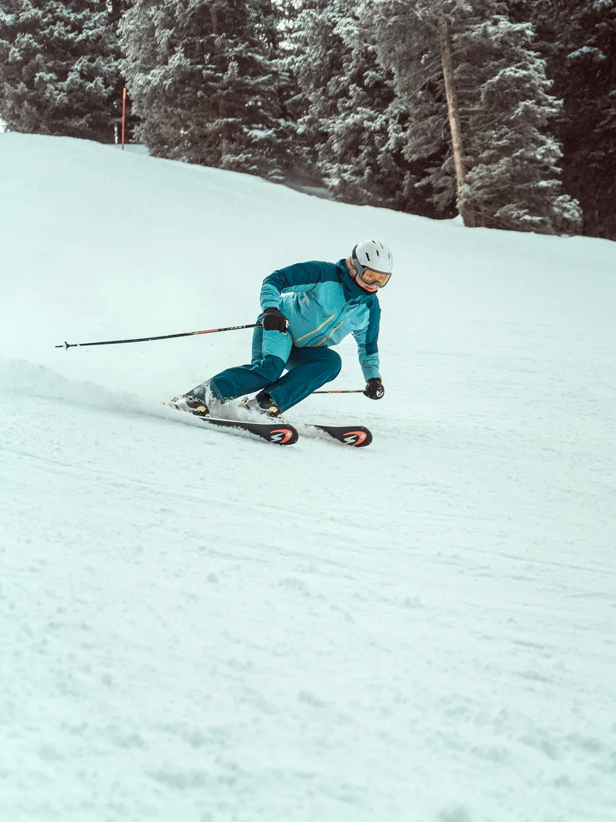 Un skieur casqué en veste turquoise descend une piste damée en lisière de forêt
