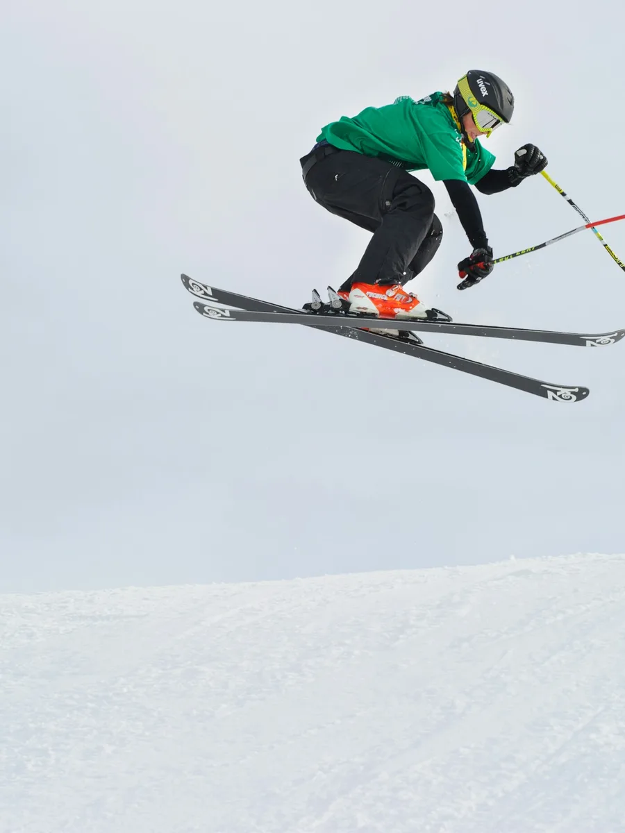 Un skieur en veste verte saute dans les airs sur fond de ciel gris.