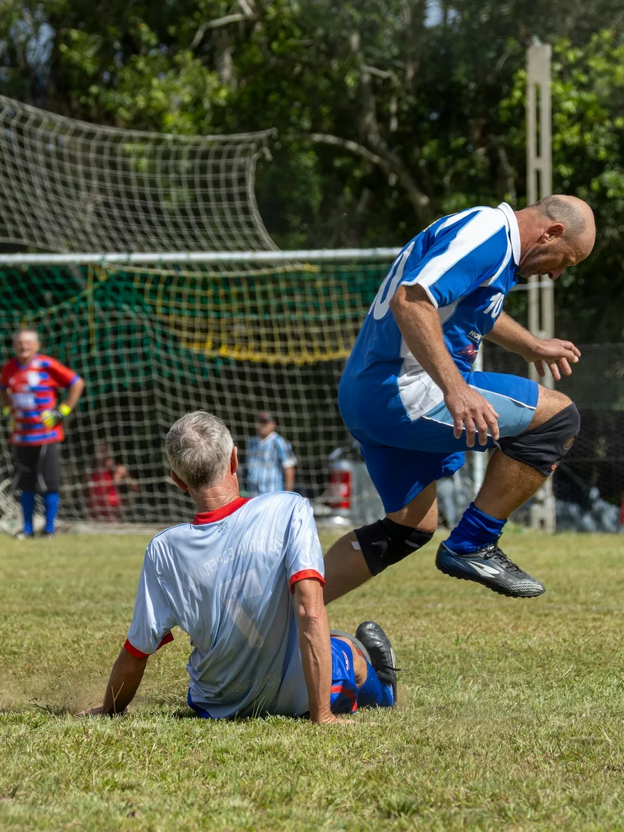 Un footballeur en maillot bleu et blanc passe au-dessus d'un adversaire tombé au sol pendant un match amateur.