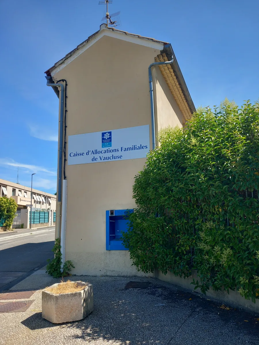 Un petit bâtiment au crépi beige portant l’enseigne de la Caisse d’allocations familiales de Vaucluse, à Carpentras, sous un ciel bleu.