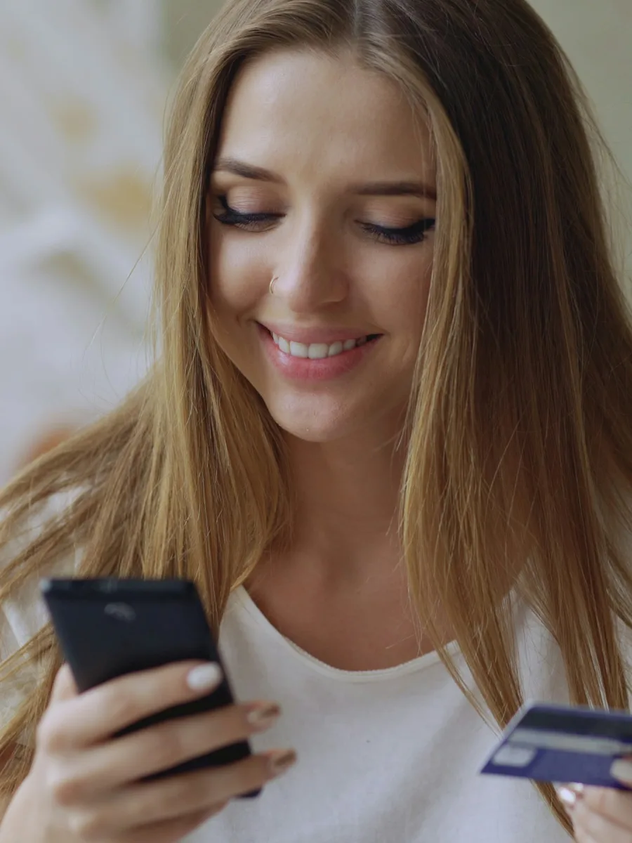 Une jeune femme souriante regarde son téléphone, une carte bancaire à la main
