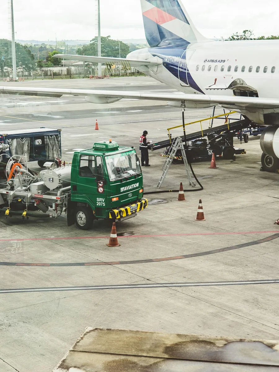 Un camion-citerne vert ravitaille un avion de ligne sur le tarmac d’un aéroport.