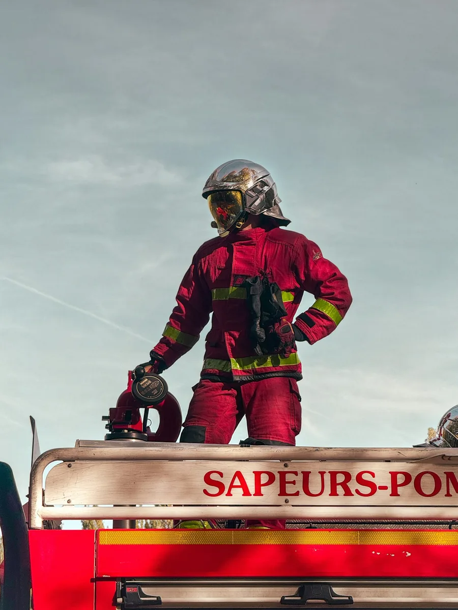 Un sapeur-pompier en tenue rouge, casque argenté, debout sur le toit d'un camion de pompiers portant l'inscription « Sapeurs-pompiers », lors d'un exercice