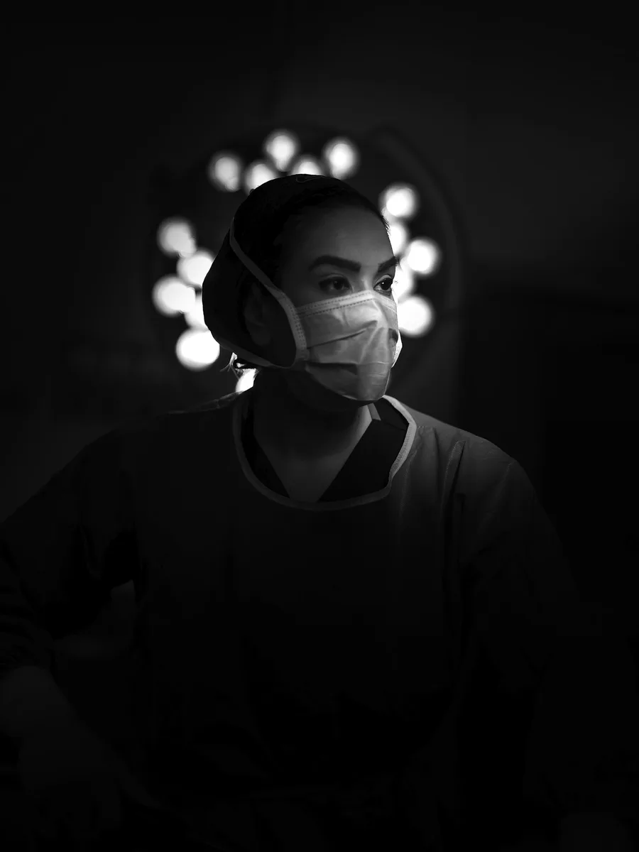 Soignante en tenue de bloc, masque et charlotte, éclairée par une lampe opératoire dans une salle sombre, photo en noir et blanc