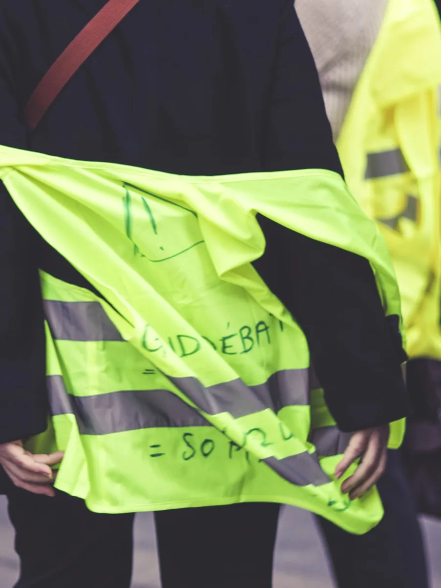 Des manifestants en gilets jaunes vus de dos, l'un portant une inscription au feutre sur son gilet