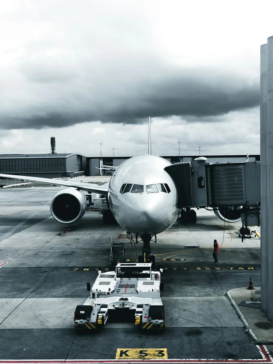 Un avion de ligne stationné à une porte d'embarquement, sous un ciel nuageux, en noir et blanc.