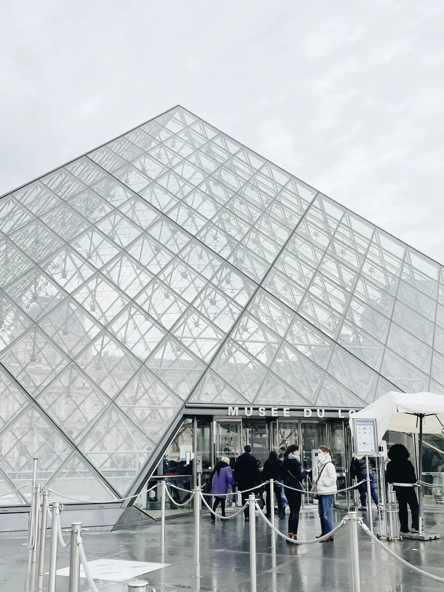La pyramide du Louvre sous un ciel gris, des visiteurs font la queue devant l'entrée