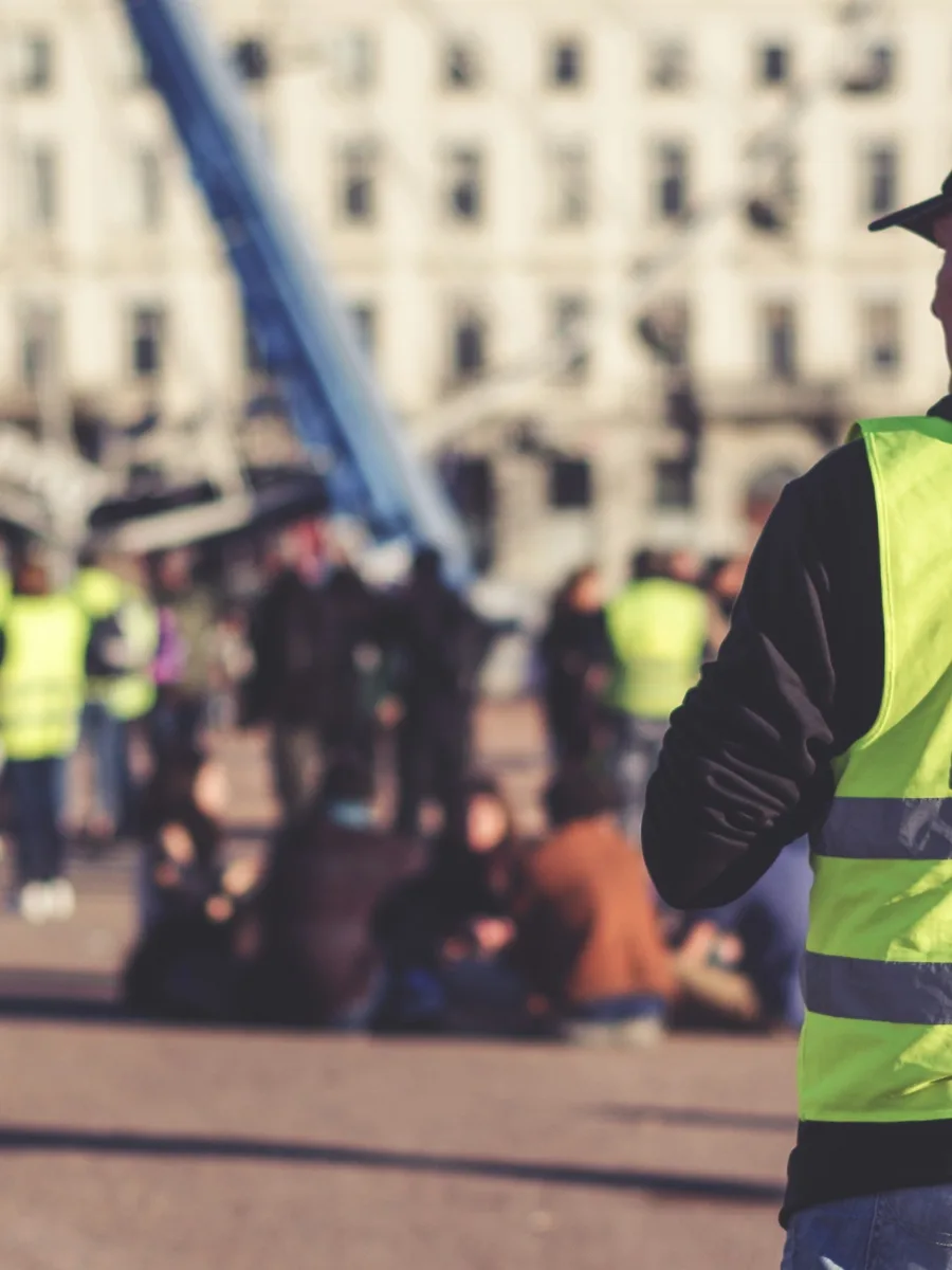Un manifestant en gilet jaune, vu de dos, sur une place où d'autres gilets jaunes sont rassemblés.