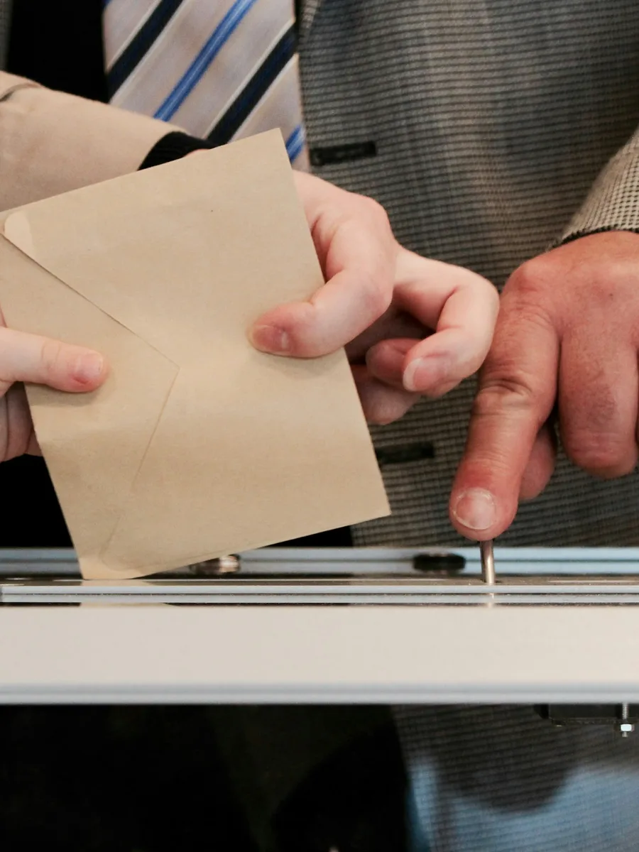 Une main glisse une enveloppe de vote dans une urne transparente lors d'une élection présidentielle française