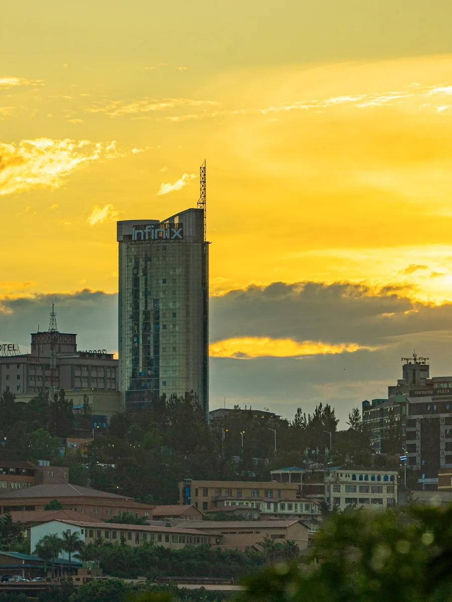 Tour vitrée dominant le centre de Kigali au coucher du soleil, sous un ciel jaune orangé, avec des immeubles et des arbres au premier plan.
