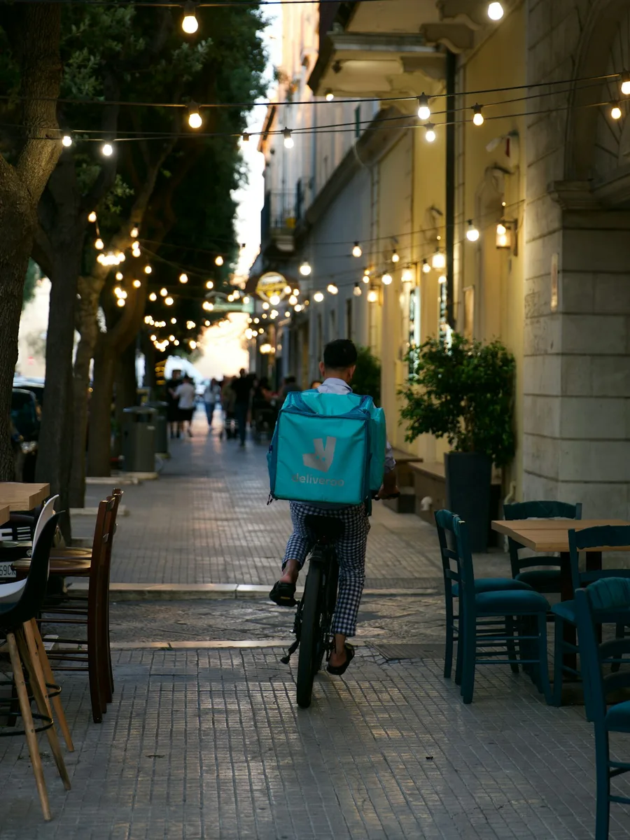 Un livreur à vélo, sac isotherme turquoise sur le dos, roule dans une rue piétonne éclairée de guirlandes