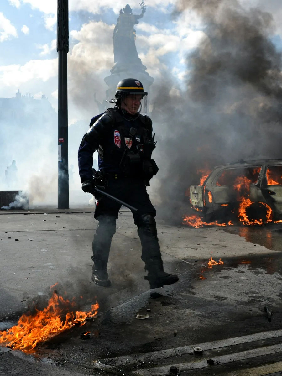 Un policier antiémeute casqué marche devant une voiture en feu et un épais nuage de fumée, place de la République à Paris