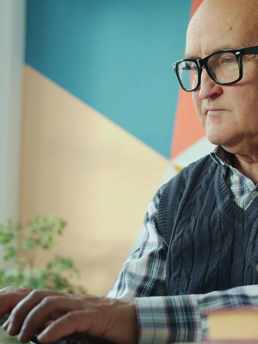 Un homme âgé à lunettes, concentré, assis devant un ordinateur portable