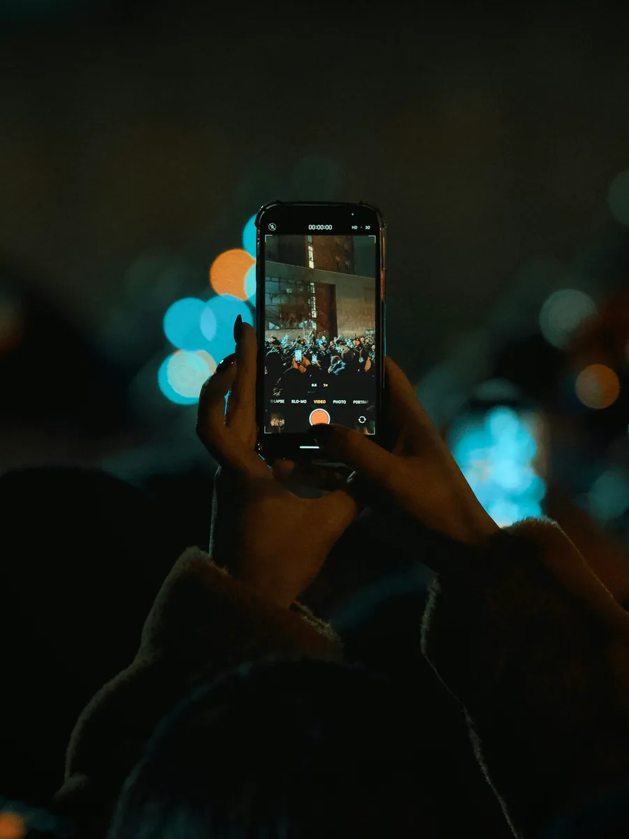 Une main tient un smartphone qui filme un rassemblement de nuit, dans une foule