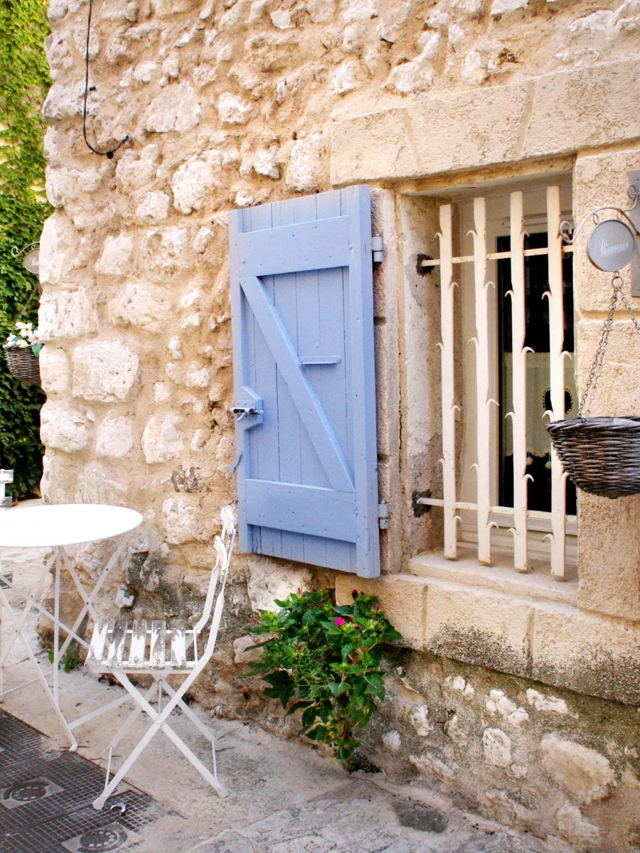 Façade en pierre d'une ruelle de village provençal, avec un volet bleu, une fenêtre grillagée et une petite table de bistrot