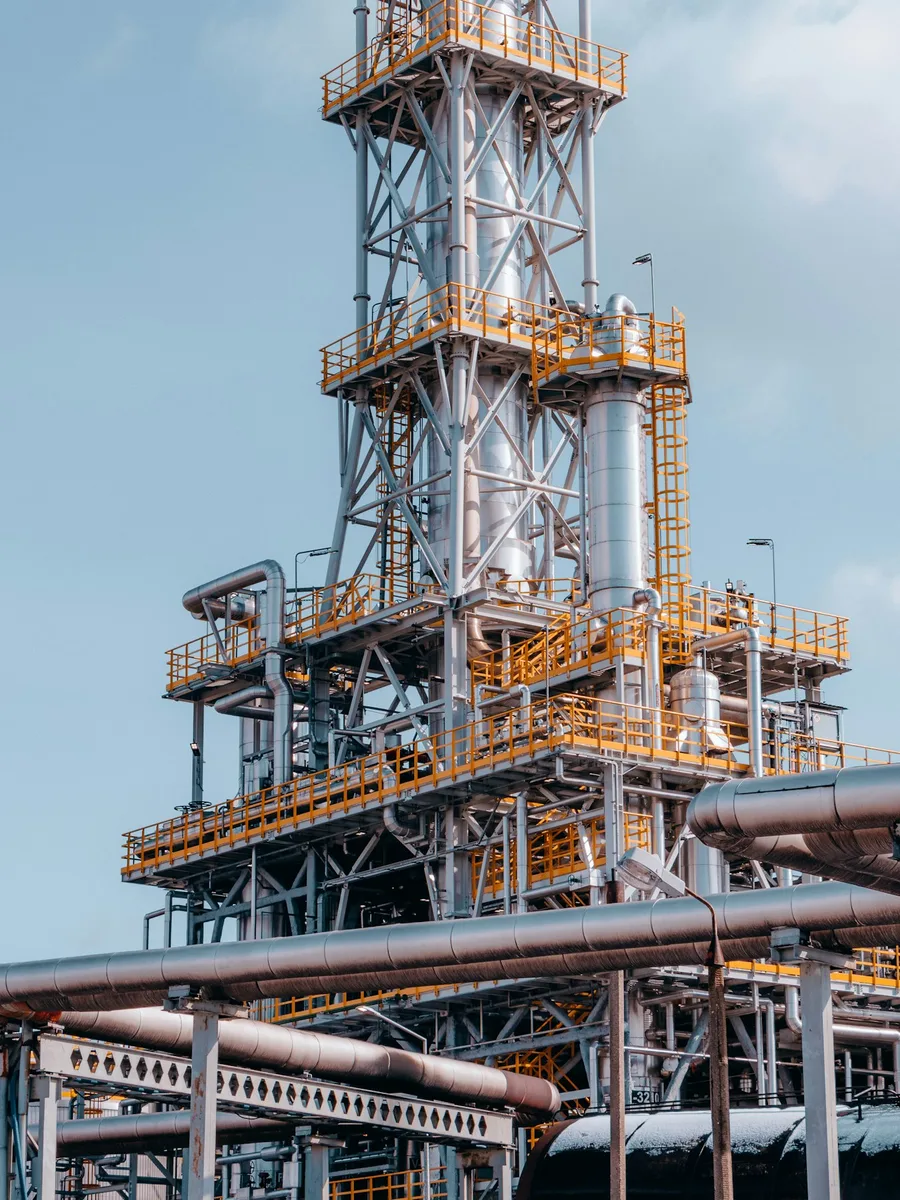 Tour métallique d'une raffinerie de pétrole, avec ses passerelles jaunes et ses canalisations, sous un ciel bleu.