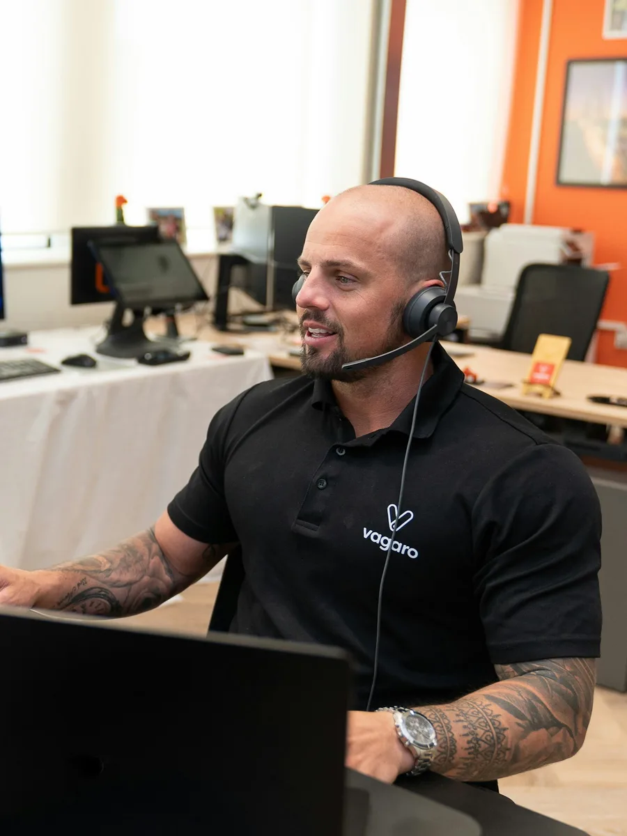 Un conseiller de service client souriant, casque sur les oreilles, devant deux écrans dans un bureau