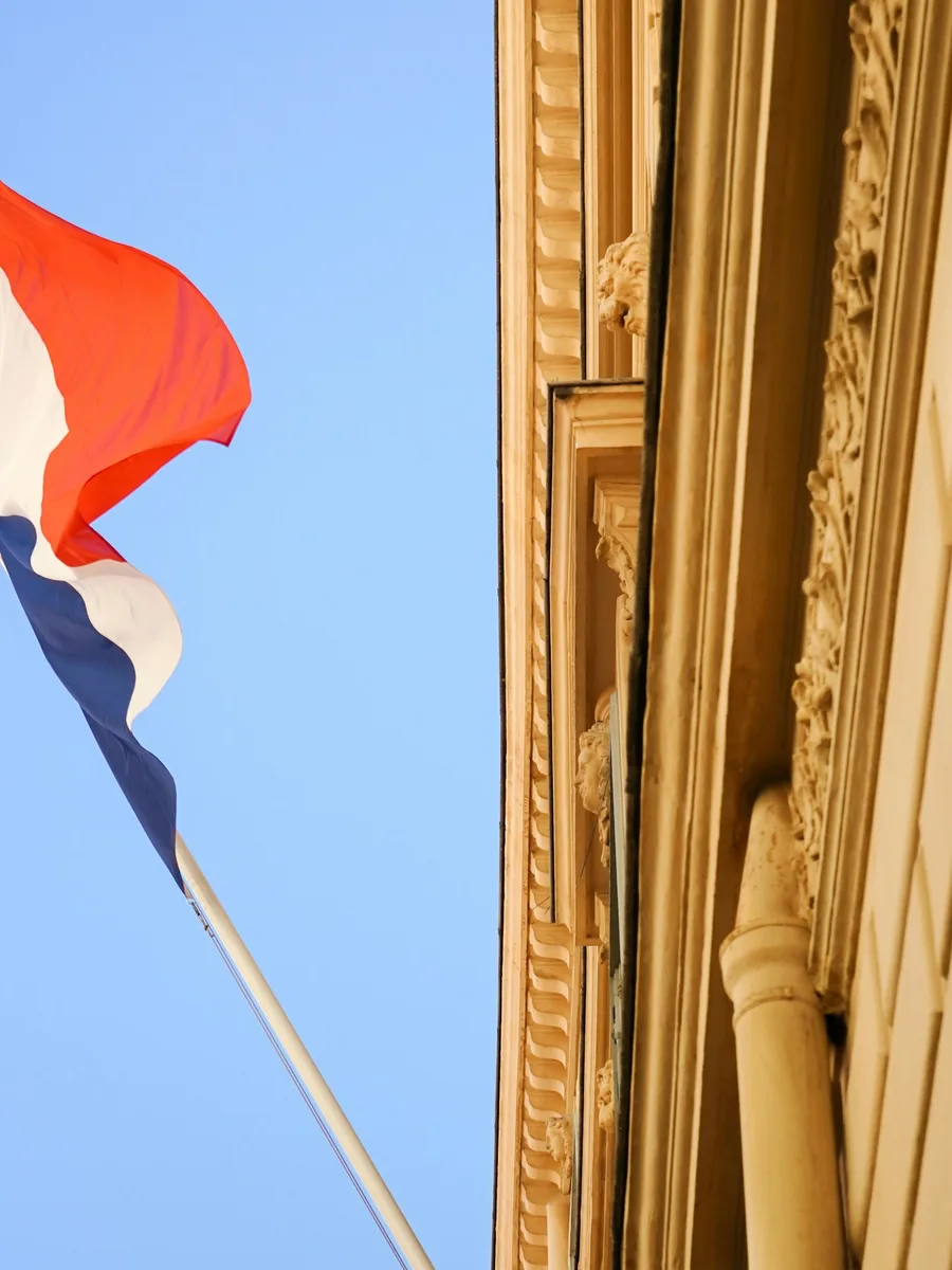 Un drapeau tricolore flotte sous un ciel bleu, à côté de la façade sculptée d'un bâtiment officiel