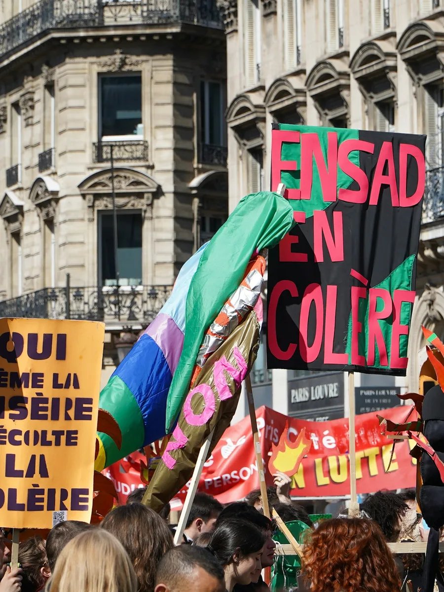 Pancartes et drapeaux dans un cortège parisien, dont une pancarte « Qui sème la misère récolte la colère », devant des immeubles haussmanniens