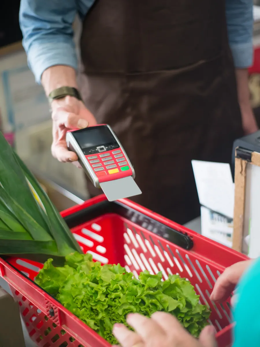 Un caissier en tablier tend un terminal de paiement au-dessus d'un panier rouge rempli de légumes