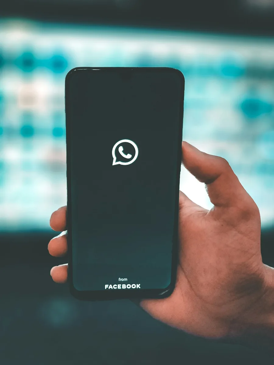 Main tenant un smartphone dont l’écran noir affiche le logo de la messagerie WhatsApp, devant un arrière-plan flou bleu-vert.