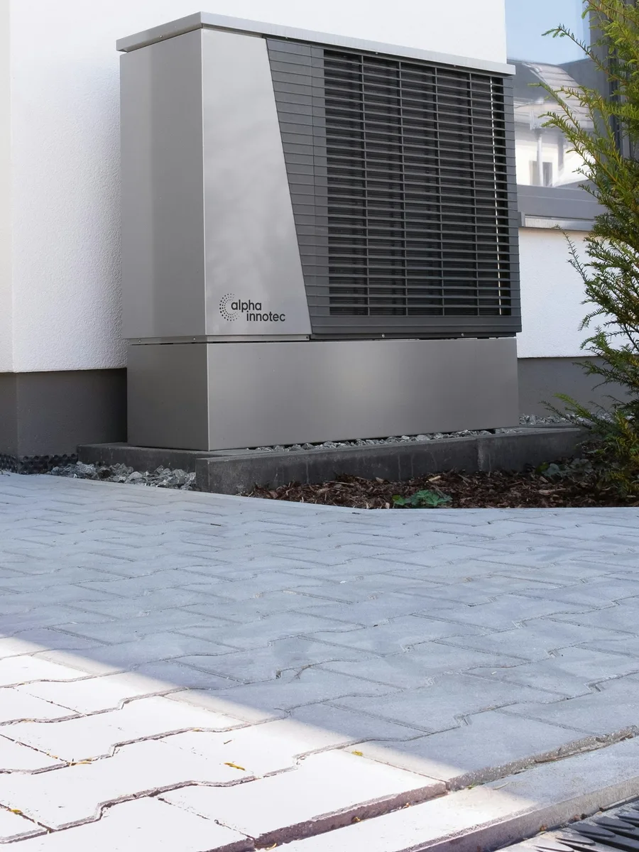 Pompe à chaleur air/eau grise installée contre la façade blanche d’une maison, au-dessus d’une allée pavée.