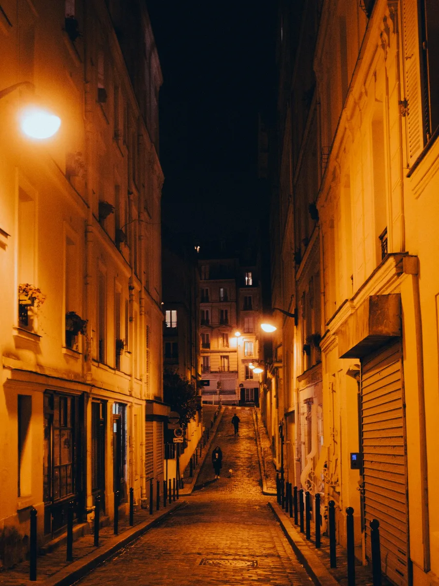 Une rue étroite de Montmartre la nuit, rideaux de fer baissés, éclairée par un lampadaire