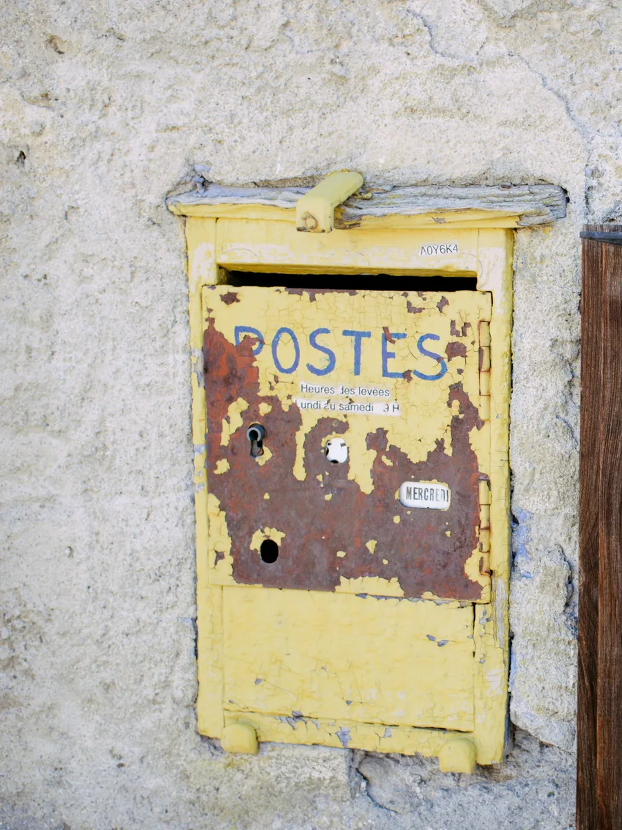 Une vieille boîte aux lettres jaune marquée « Postes » sur un mur de pierre, à côté d'un volet en bois