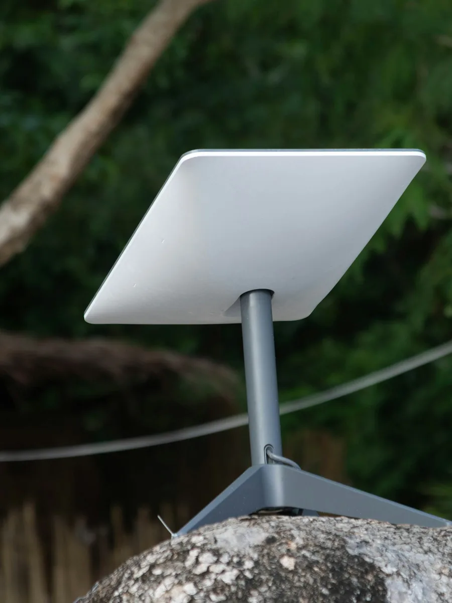 Une antenne satellite plate et blanche sur son pied, posée sur un rocher devant la végétation.