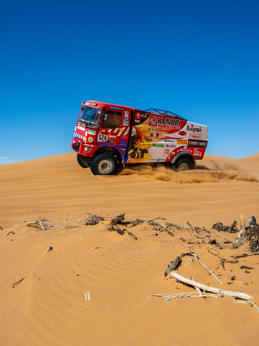 Un camion de rallye-raid rouge franchit une dune de sable sous un ciel bleu