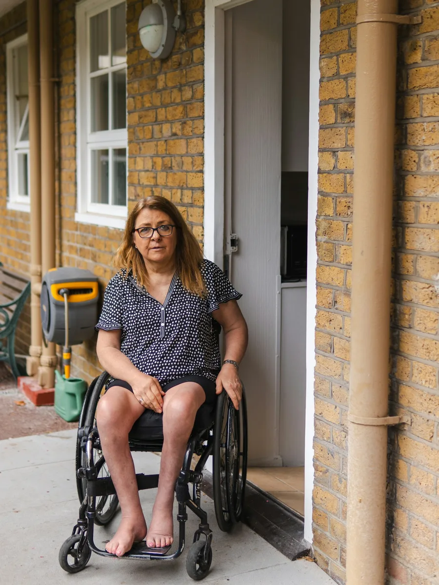 Une femme en fauteuil roulant devant la porte de son logement, dans une cour en briques.