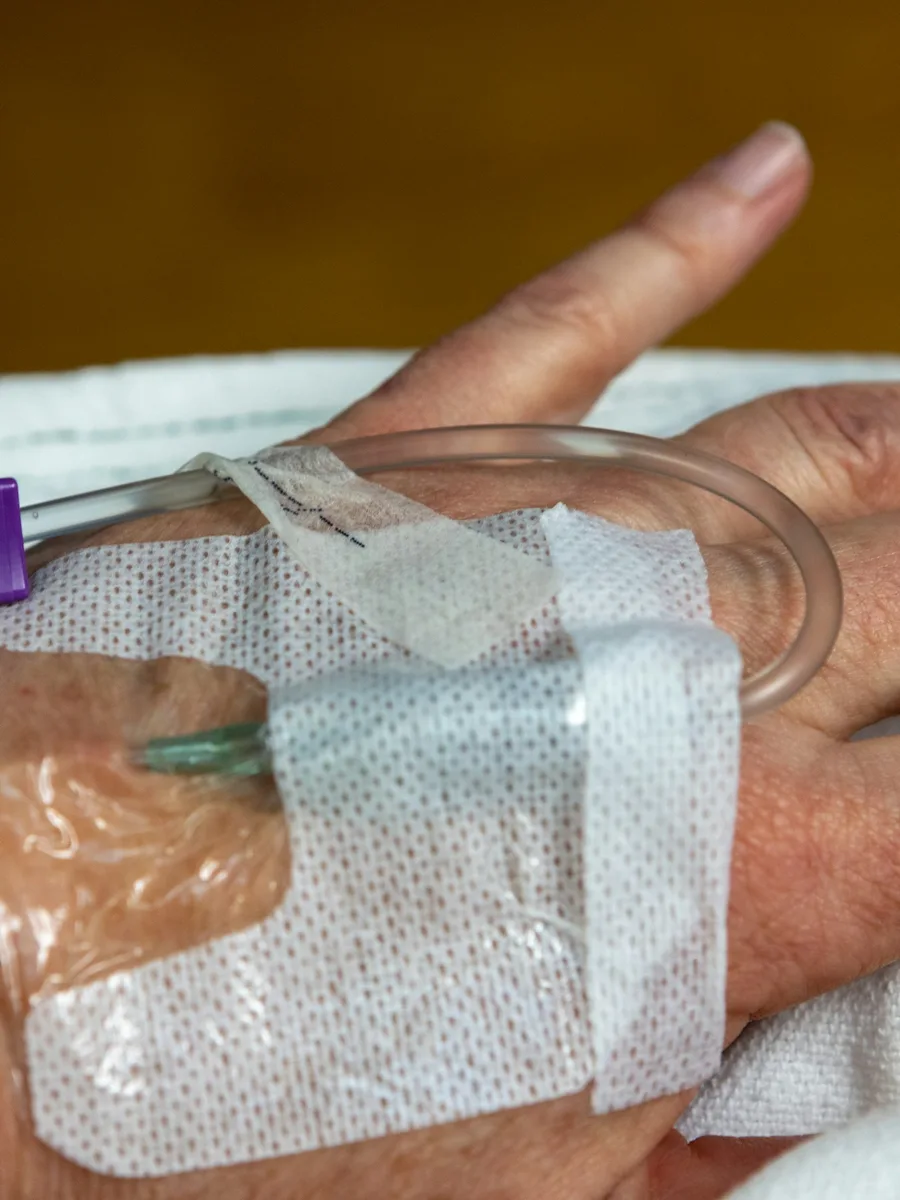 Une main de patient allongé, avec un cathéter de perfusion maintenu par un pansement transparent