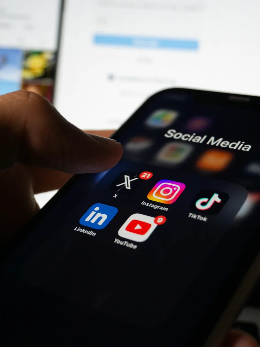 Une main tient un smartphone dont l'écran affiche un dossier « Social Media » avec les icônes de X, Instagram, TikTok, LinkedIn et YouTube